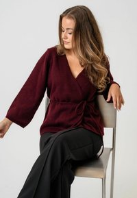 Pulover burgundy cu maneci lungi, dintr-un material tricotat texturat, cu talie încrucișată și decolteu în V, purtat împreună cu pantaloni negri.