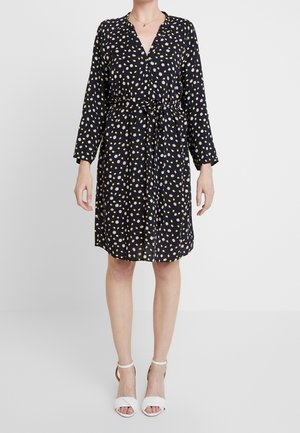 Frau trägt ein knielanges schwarzes Kleid mit weiß-gelbem Blumenmuster, langen Ärmeln, einem Taillengürtel und weißen offenen Sandalen.