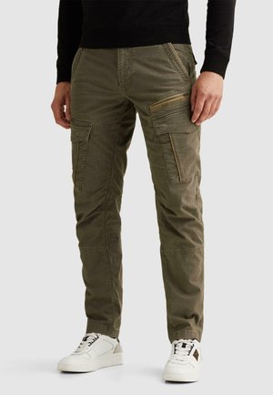 Uomo che indossa pantaloni cargo verde oliva con molte tasche e sneaker bianche, in piedi contro uno sfondo semplice.