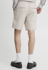 Beige bomullsshorts med elastisk midje, to glidelås-lommer bak og oppbrettede sømmer. Brukt med hvite sokker og mørke sko.