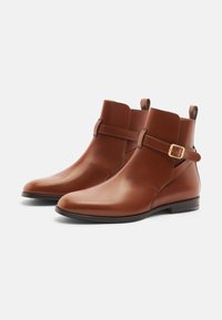 Braune Leder-Stiefeletten mit runder Zehenform, flachem Absatz, seitlichem Schnallen-Detail und kurzem elastischem Einsatz auf einer glatten Oberfläche.