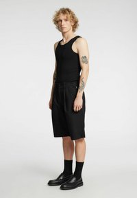 Czarny top na ramiączkach, czarne szorty i czarne mokasyny. Widoczne tatuaże na ramionach; model stoi z rękami luźno opuszczonymi wzdłuż ciała. Prosty design.