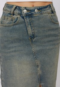 Jupe en denim bleu clair avec une fente à l'avant, deux boutons à la taille, des poches et des détails de couture contrastés. Texture lisse.