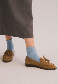 Hnědé semišové mokasíny s detailem mašle, spárovány s žebrovanými světle modrými ponožkami, nošeny s šedou sukní, ukazující jednoduchý design a barevný kontrast.