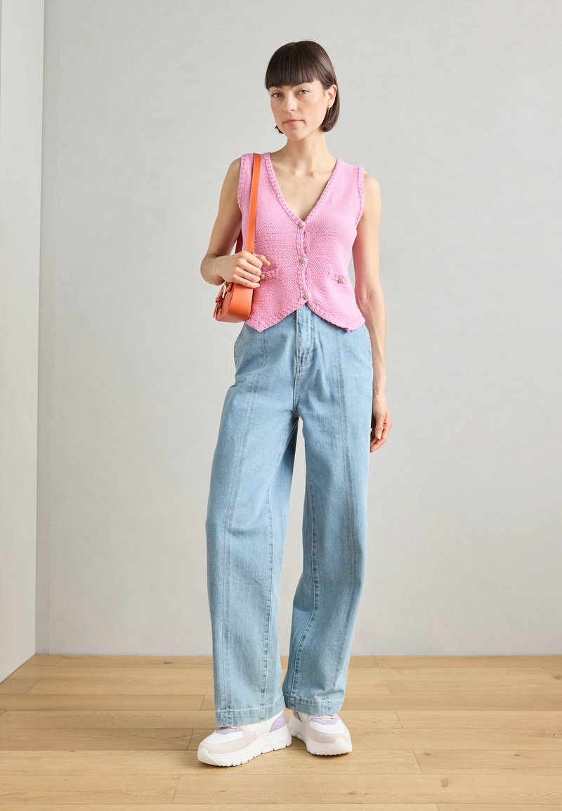 Rosa gestrickte Weste mit V-Ausschnitt und Knopfdetails, kombiniert mit hellblauen weit geschnittenen Jeans und weißen Plateausneakern. Orangefarbene Umhängetasche.