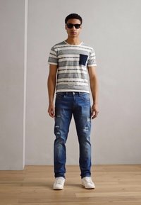 Camiseta a rayas con bolsillo azul marino, combinada con unos vaqueros azules desgastados y zapatillas blancas. Modelo lleva gafas de sol negras y se encuentra de pie contra una pared clara.