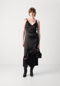 Schwarzes Satinslipkleid mit Spitzenbesatz, verstellbaren Trägern und V-Ausschnitt. Das Model trägt knöchelhohe schwarze Stiefel und eine kleine Tasche.