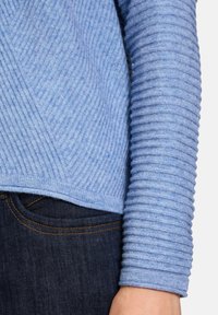 Hellblauer gestrickter Pullover mit diagonaler Rippenstruktur am Körper und vertikaler Rippenstruktur an den Ärmeln, kombiniert mit dunklen Jeans.