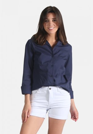Chemise boutonnée bleu marine à manches retroussées, en tissu lisse, associée à un short en denim blanc. La chemise présente un col classique.