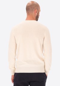 Uomo con testa rasata e barba, indossa un maglione a maniche lunghe color crema e pantaloni neri, visto di spalle su uno sfondo neutro.