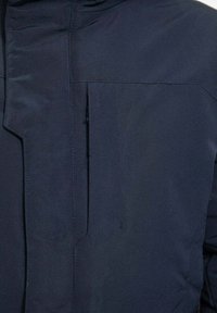 Veste bleu marine avec un tissu lisse et texturé. Comprend une poche poitrine zippée et des coutures discrètes le long des coutures.