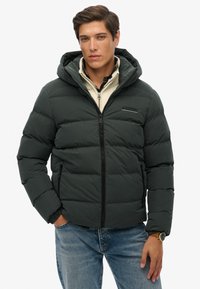 Chaqueta acolchada verde oscura con capucha, cierre frontal y bolsillos laterales. Presenta un diseño acolchado y una marca en el pecho izquierdo. Llevada sobre un suéter ligero.