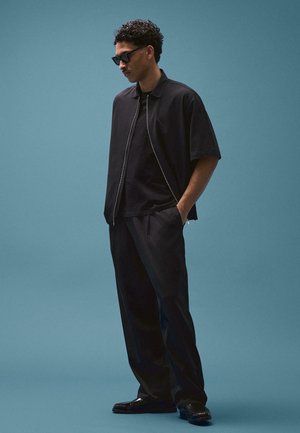 Muškarac u crnim sunčanim naočalama, crnoj jakni kratkih rukava sa patentnim zatvaračem, crnoj majici, crnim hlačama i crnim cipelama, stoji na plavoj pozadini.