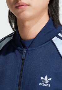 Námořnická zipová bunda sportovního oblečení ze strukturované látky, s vysokým límcem, světlemodrými akcentovými pruhy a bílým vyšívaným logem.