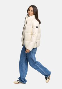 Giacca puffer avorio con colletto alto, zip frontale e patch con logo nero, abbinata a jeans blu larghi e sneakers crema con strisce nere.