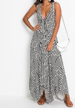 Femme portant une robe maxi sans manches à motif géométrique noir et blanc, avec un profond décolleté en V et un sac blanc à l'épaule.