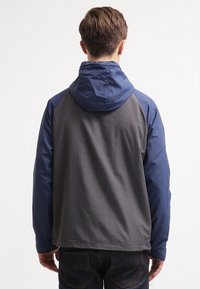 Mand iført en grå og blå hættejakke, vendt væk, med mørkeblå jeans synlige mod en ensfarvet hvid baggrund.