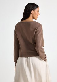 Pull en tricot marron avec un grand col, ourlet côtelé et épaules tombantes. Présente une texture lisse et une coupe décontractée, associé à une jupe légère.