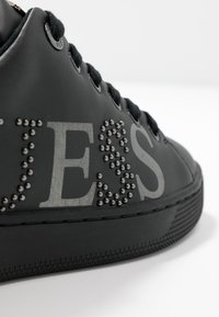 Zwarte sneaker met een gladde afwerking, voorzien van metallic studs en gedurfde grijze letters. Een gestructureerde zool en zwarte veters maken het ontwerp af.