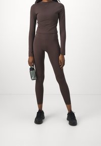 Maglia marrone a maniche lunghe e leggings abbinati in materiale elasticizzato, accompagnati da una bottiglia d'acqua in mano e scarpe da ginnastica nere.