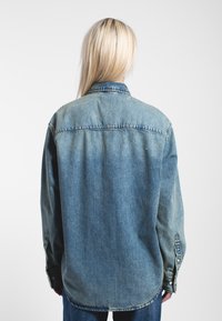 Camicia di jeans, colore azzurro chiaro, vestibilità rilassata, maniche lunghe con polsini a bottone, cuciture visibili lungo le cuciture, orlo curvo.