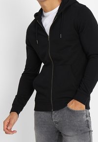 Felpa nera con zip, realizzata in tessuto morbido, con tasca frontale, cappuccio regolabile con coulisse e polsini a costine.