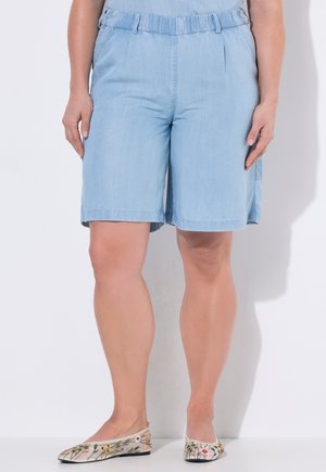 Vrouw draagt lichtblauwe knielange korte broek en platte schoenen met bloemenpatroon, staand tegen een witte achtergrond.