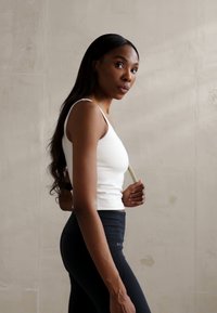 Hvit ermeløs topp og svarte høyt liv leggings, med en glatt tekstur og tettsittende silhuett. Bakgrunn av lys betong.