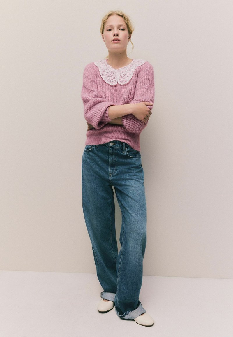 Rosa Strickpullover mit einem weißen Spitzenkragen, kombiniert mit lockeren blauen Denim-Jeans und cremefarbenen Schuhen. Texturiertes Material und lockere Passform.