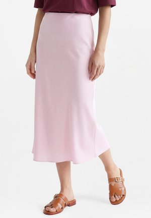 Frau trägt einen hoch taillierten, hellrosa Midirock, kombiniert mit braunen Ledersandalen, vor einem schlichten Hintergrund.