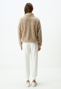 Beige pluchen pullover met een hoge kraag en lange mouwen, gecombineerd met witte broek met omslag en lichte sneakers, gezien vanaf de achterkant.