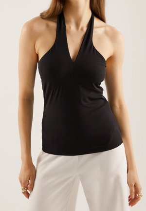 Femme portant un haut dos-nu noir et un pantalon blanc, avec des bagues aux deux mains sur un fond neutre.