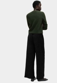 Ciemnozielony sweter o teksturowanym wykończeniu, zestawiony z szerokimi czarnymi spodniami z pionowym prążkiem i czarnymi płaskimi butami.
