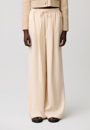 Persona con pantalones beige de pierna ancha con cintura de cordón y chaqueta corta beige, de pie frente a un fondo claro y liso.