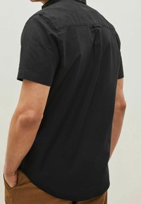Chemise noire à manches courtes en coton, avec un pli dans le dos et un ourlet arrondi. Texture lisse avec une coupe décontractée.