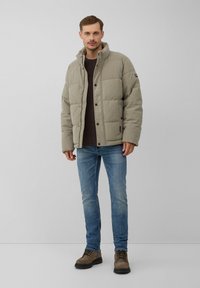 Leichte, gepolsterte Jacke in Hellgrau mit hohem Kragen und Druckknöpfen. Getragen über einem gestreiften Hemd, kombiniert mit blauen Jeans und braunen Stiefeln.