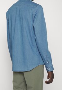 Camisa de mezclilla de manga larga en azul claro con un yugo en la espalda, puños abotonados y detalles de costura sutiles, combinada con pantalones verdes.