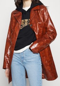 Abrigo trench de imitación de cuero marrón brillante con cuello de muesca, cierre de un botón y bolsillos laterales, llevado sobre una sudadera negra con capucha y jeans.