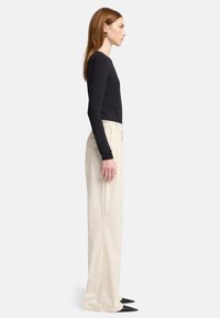 Zwarte lange mouw top gecombineerd met loszittende beige broek; model draagt zwarte puntige schoenen. Zijaanzicht dat de pasvorm en kleurcontrast benadrukt.