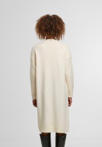 Krémový cardigan do délky kolen, vyrobený z měkké, texturované látky, s dlouhými rukávy a žebrovaným lemem. Bez viditelných zapínání nebo vzorů.