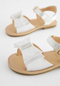 Vita sandaler med en glansig satindubbelrosett, öppen tå-design, läderliknande sula och justerbar rem med ett guldspänne.