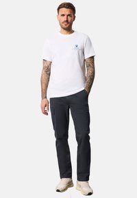 Camiseta blanca de algodón con gráfico azul y pantalones cargo oscuros. Zapatillas gris claro complementan el conjunto. Tatuajes visibles en los brazos.