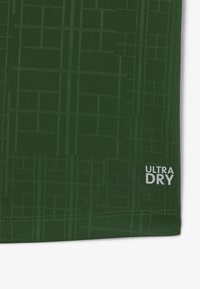 Tessuto verde scuro con un sottile motivo a griglia e testo bianco "ULTRA DRY" vicino all'orlo cucito nell'angolo in basso a destra.