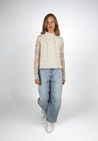 Frau, die auf einem weißen Hintergrund in einem weißen, gestrickten, ärmellosen Pullover, einem gemusterten, durchsichtigen Langarmoberteil, lockeren, hellblauen Jeans und weißen Turnschuhen nach vorne geht.