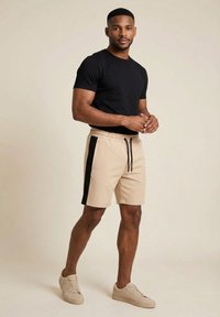 Hombre de pie con una camiseta negra de manga corta, pantalones cortos beige con franjas negras laterales y zapatillas beige sobre un fondo sencillo.
