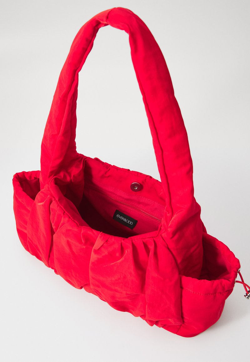 Bolso acolchado rojo con una textura suave, con un solo asa acolchada y un bolsillo interior con cremallera. Cierre de cuerda en un lado.