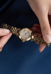 Orologio dorato con bracciale testurizzato, quadrante rotondo bianco, numeri romani e accenti di diamante. Le lancette sono impostate su un'ora specifica.