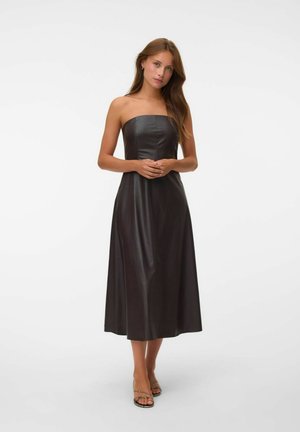 Joven mujer con cabello largo y castaño que lleva un vestido midi de cuero negro sin tirantes y tacones de tiras, de pie con las manos entrelazadas.