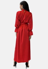 Robe maxi en satin rouge avec manches longues bouffantes, col montant et un détail dos en forme de goutte. Une ceinture à la taille met en valeur la silhouette.