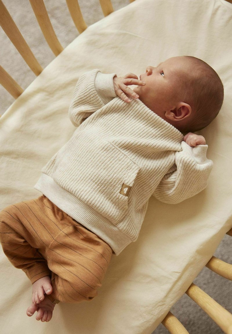 Maglione a coste crema con tasca e pantaloni a righe marroni, posati su una superficie di tessuto chiaro, che mostrano il bambino in una posizione comoda.
